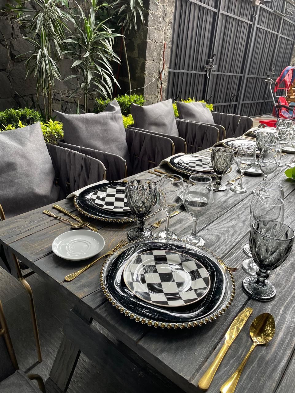 Mesa de comedor formal al aire libre con platos a cuadros, cubiertos dorados y sillas grises.