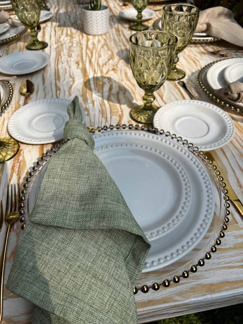 Mesa con platos blancos, detalles dorados, vasos verdes y servilleta anudada.