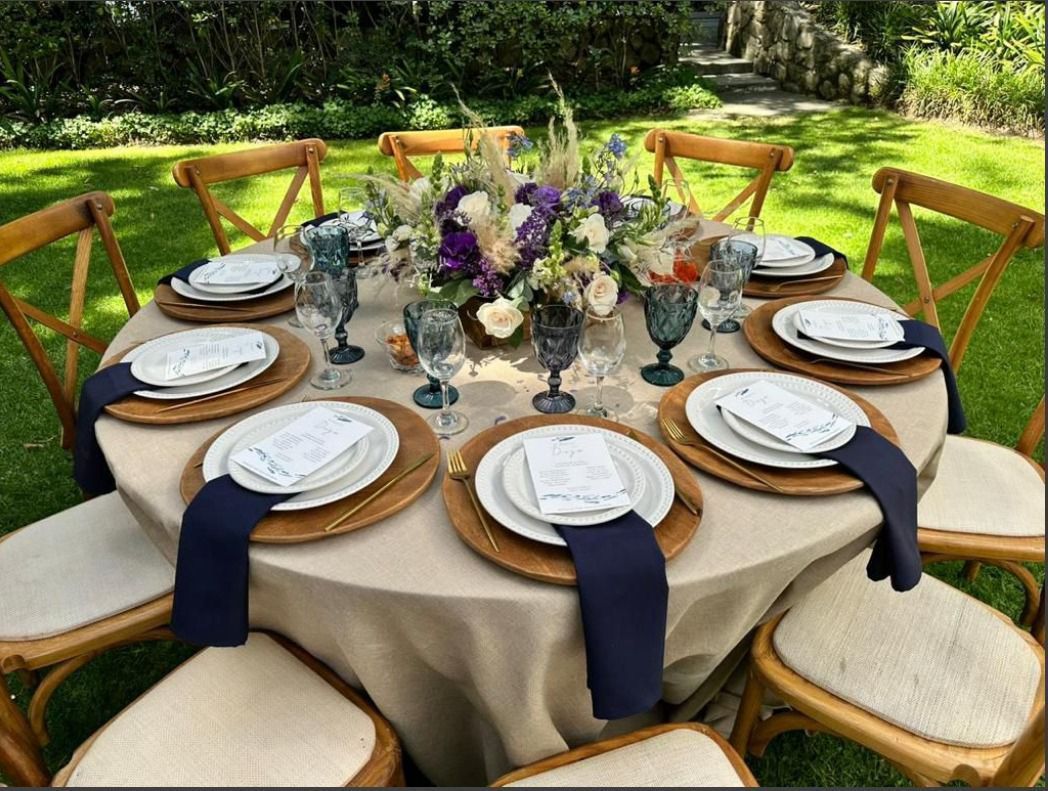 Mesa redonda para comer al aire libre, con centro floral, cristalería azul y cubiertos.