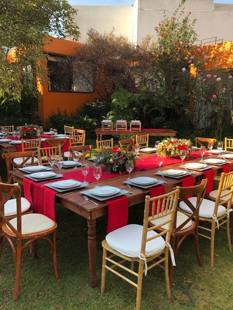 Mesas de comedor al aire libre con caminos rojos, vajilla y centros de mesa florales, rodeadas de sillas en un césped.