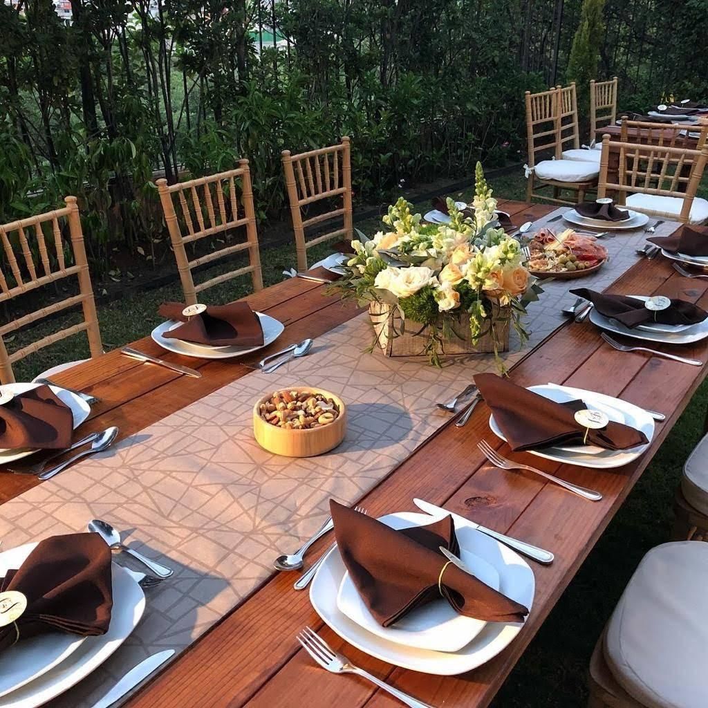 Mesa larga de madera para comer al aire libre, con centro floral, platos, servilletas y sillas.