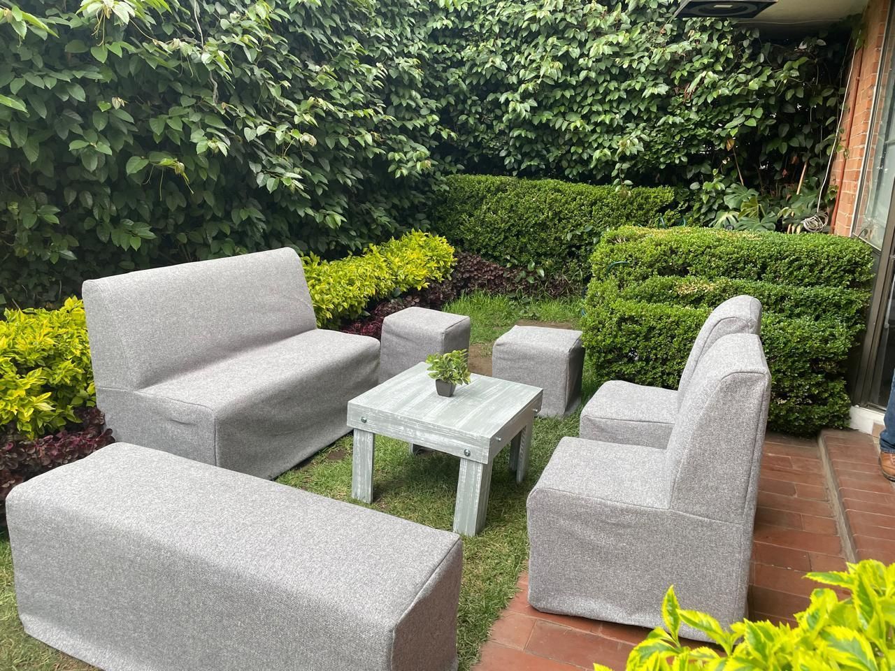 Disposición de asientos al aire libre: sofá gris, sillas y mesa en un patio, rodeado de vegetación.