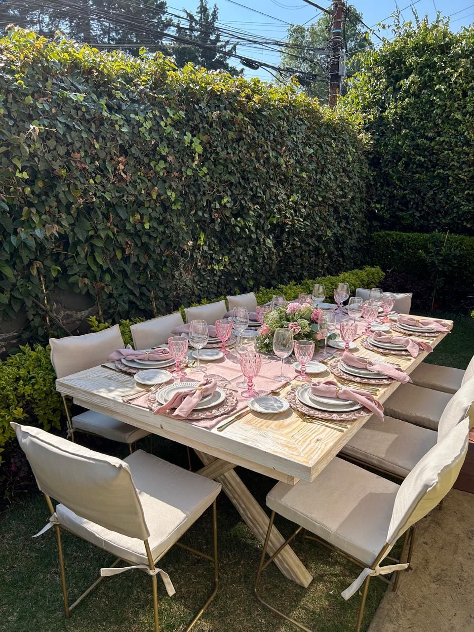 Mesa de comedor al aire libre para una reunión con detalles en rosa y sillas blancas contra un seto verde.