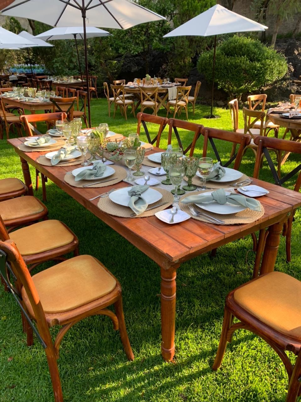 Mesas de comedor al aire libre con platos y vasos en un césped bajo sombrillas.