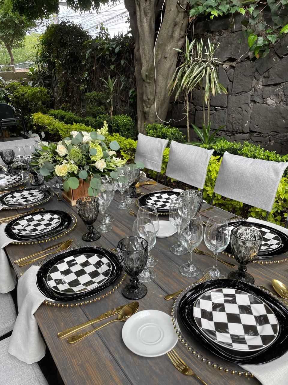 Juego de mesa de comedor formal para exteriores con platos a cuadros, detalles dorados y centro de mesa floral.