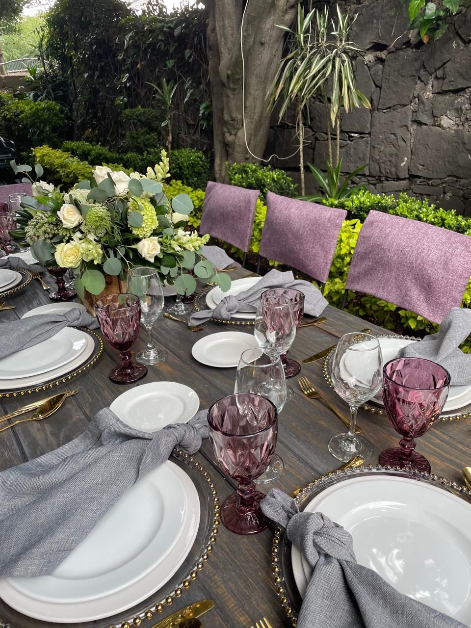 Mesa para exterior con centro de mesa floral, copas moradas y servilletas grises.
