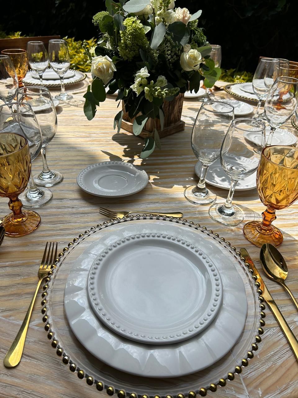 Elegante mesa para exterior con centro de mesa floral, detalles dorados y cubiertos.