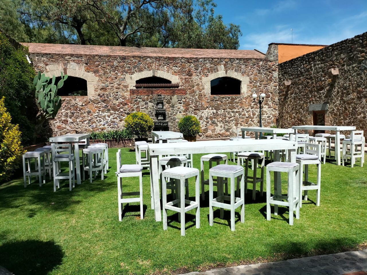 Espacio para eventos al aire libre con mesas blancas y taburetes sobre césped verde, edificio de piedra al fondo.