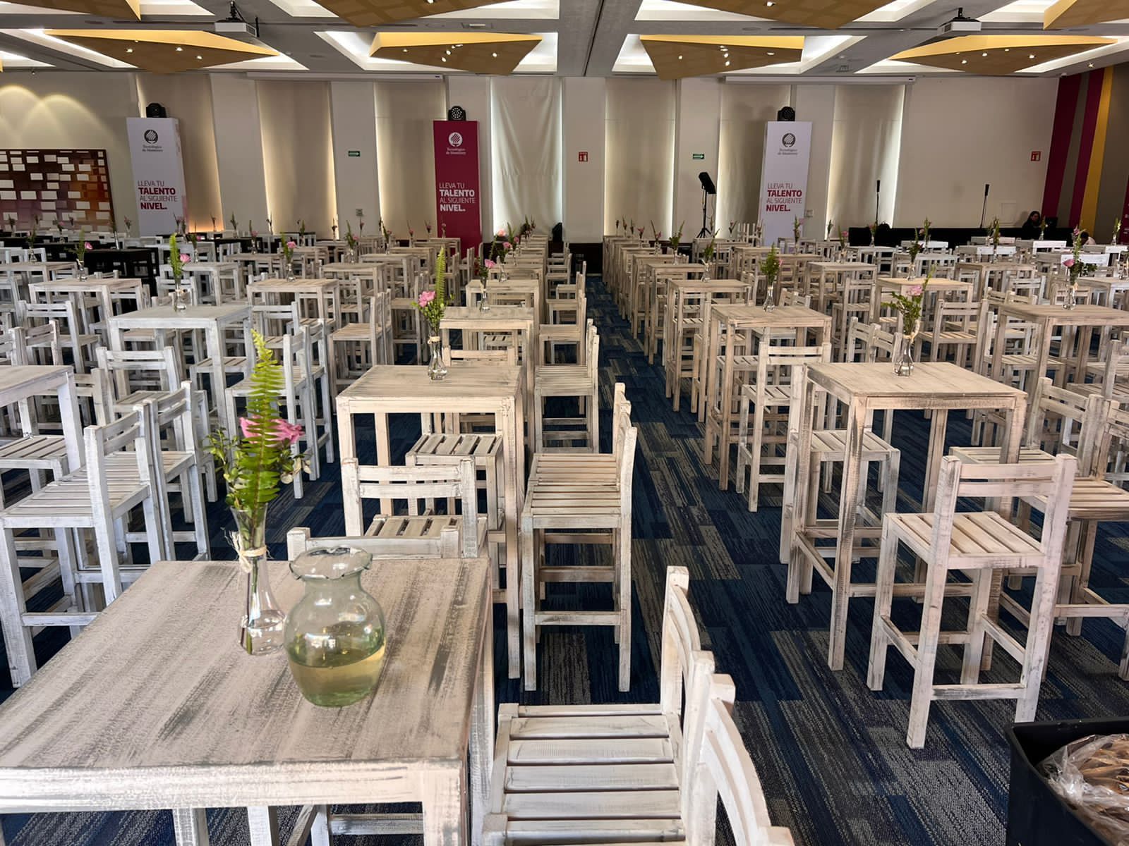 Salón de eventos vacío con filas de mesas y sillas blancas, centros de mesa florales.