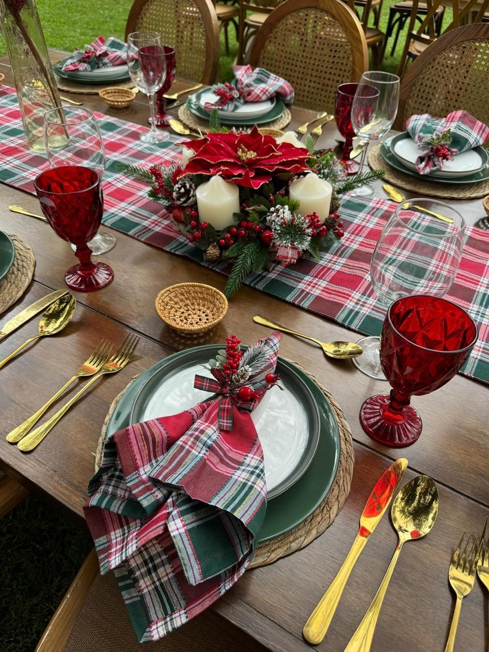 Mesa navideña con camino de mesa a cuadros, cubiertos dorados, copas rojas y centro de mesa festivo.