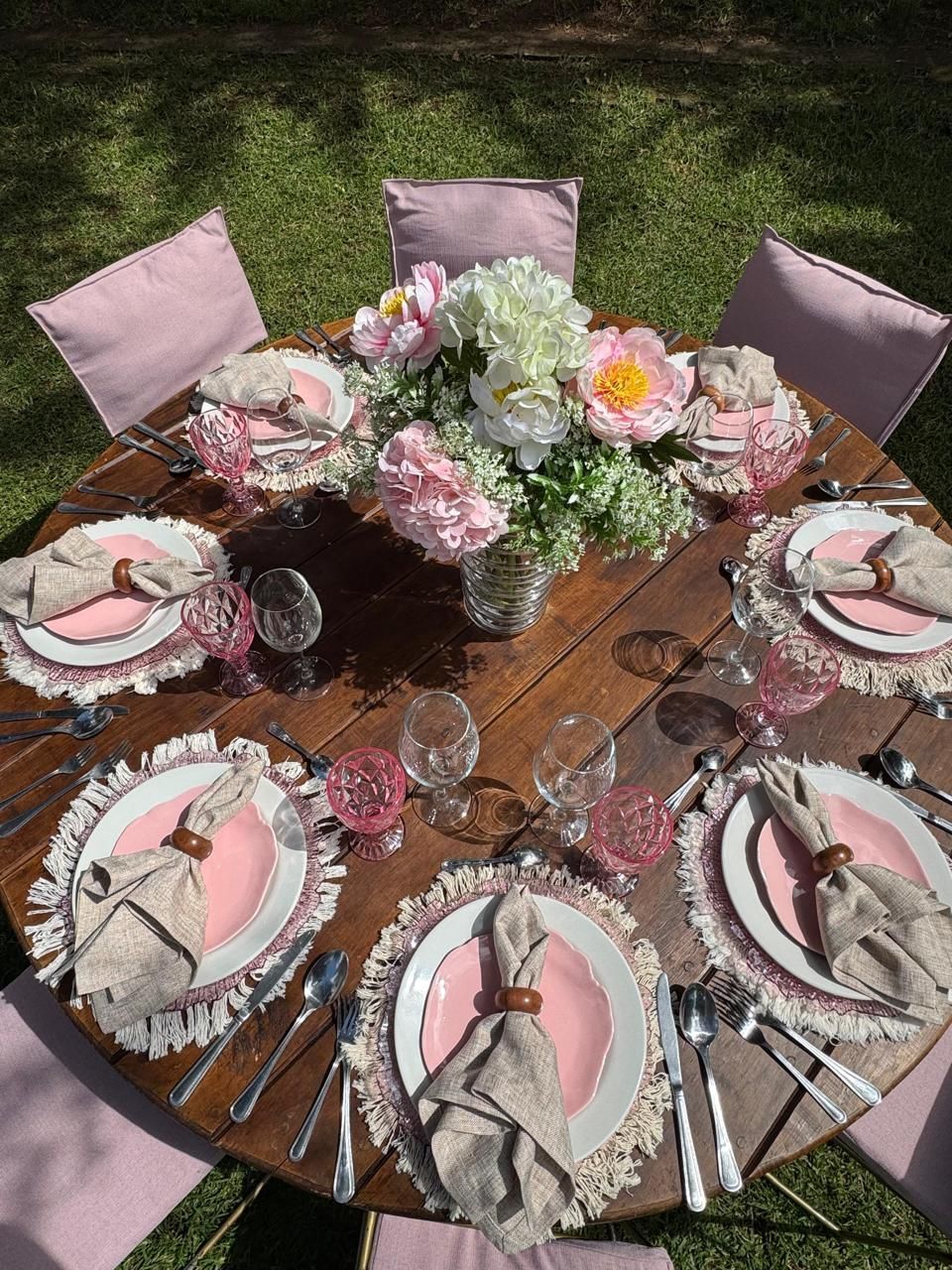 Mesa redonda de madera para picnic con platos rosas, servilletas y centro de mesa floral, al aire libre sobre césped.