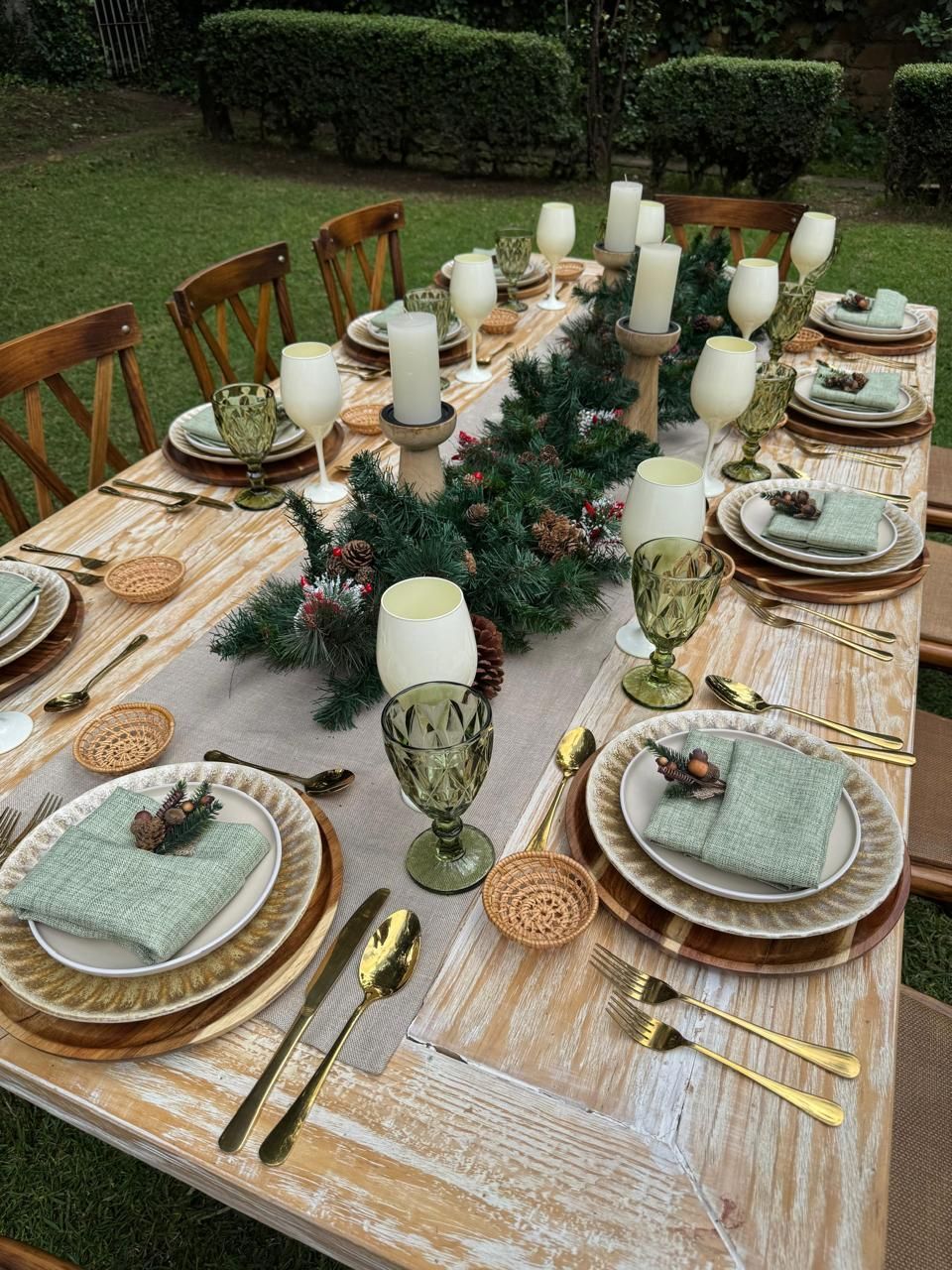 Mesa de comedor festiva al aire libre con platos con borde dorado, servilletas verdes, velas y guirnalda de hojas perennes.