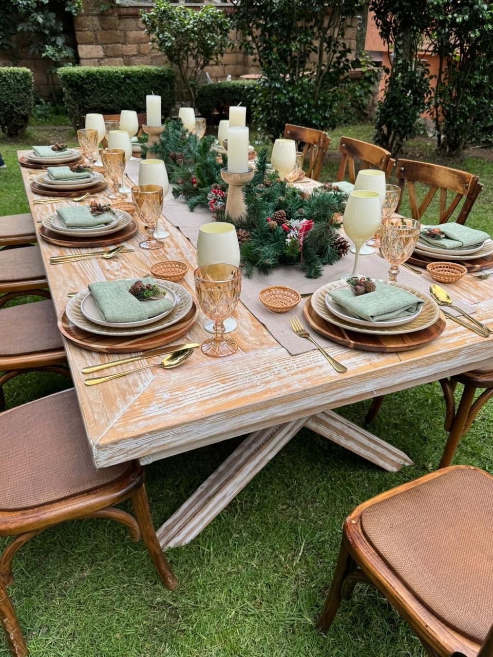 Mesa larga de madera dispuesta para comer al aire libre, decorada con velas y vegetación.