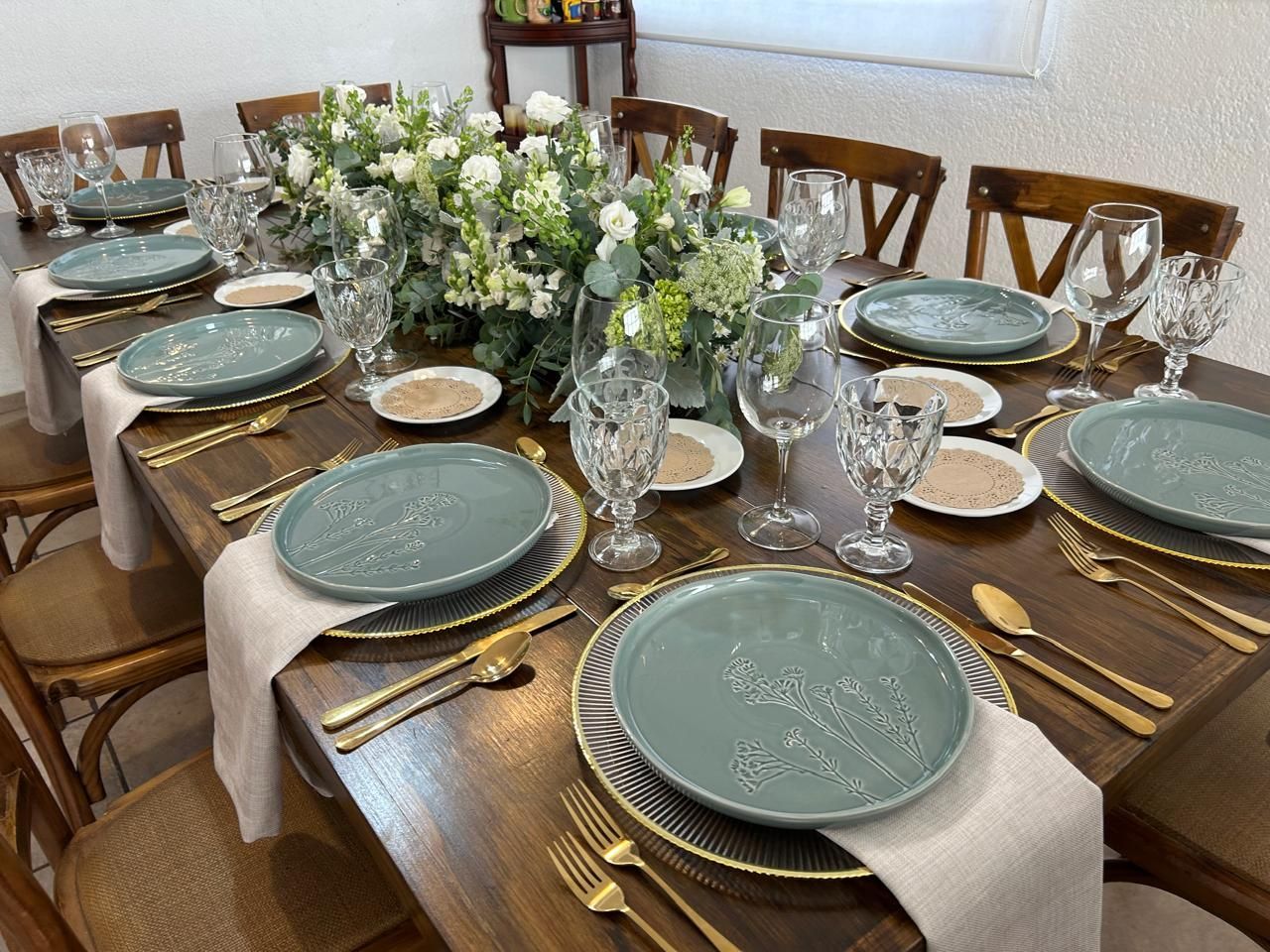 Mesa de madera puesta para comer, con centro de mesa floral, platos verdes, cubiertos dorados y vasos transparentes.