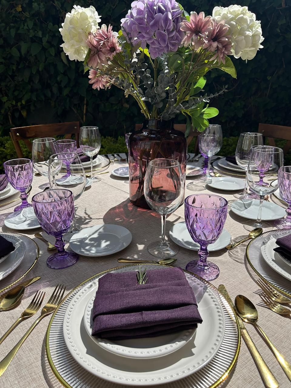 Elegante mesa con detalles en morado, con flores, vasos y cubiertos dorados al aire libre.