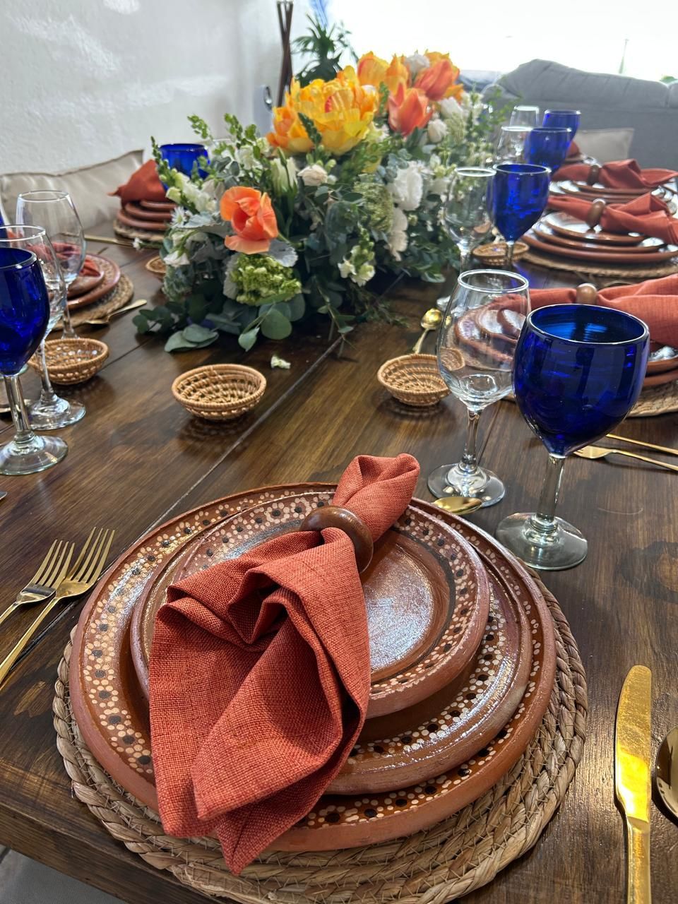 Mesa de comedor festiva con platos de terracota, servilletas de color óxido y cristalería azul, rematada con un centro de mesa floral.