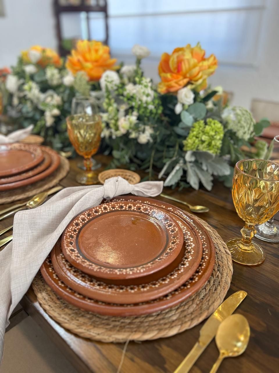 Decoración de mesa con platos de terracota, cubiertos dorados, copas de color ámbar y centro de mesa floral.