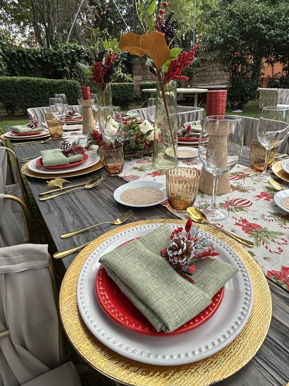 Juego de mesa de comedor al aire libre para una comida navideña con detalles dorados, platos rojos y verdes y centros de mesa florales.