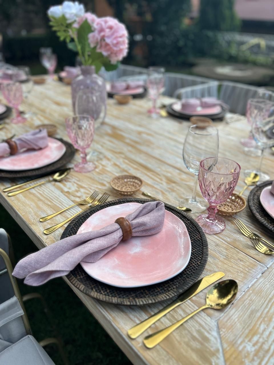 Mesa al aire libre con platos rosas, cubiertos dorados y cristalería rosa, coronada con un jarrón de flores rosas.