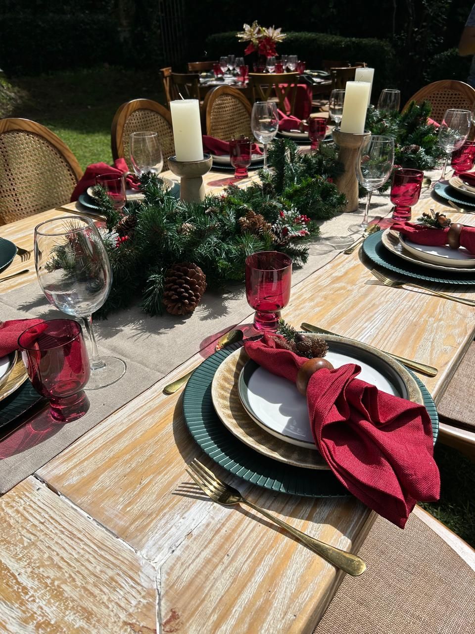 Mesa festiva al aire libre. Cubiertos verdes y rojos con centros de mesa de pino y velas sobre una mesa de madera clara.