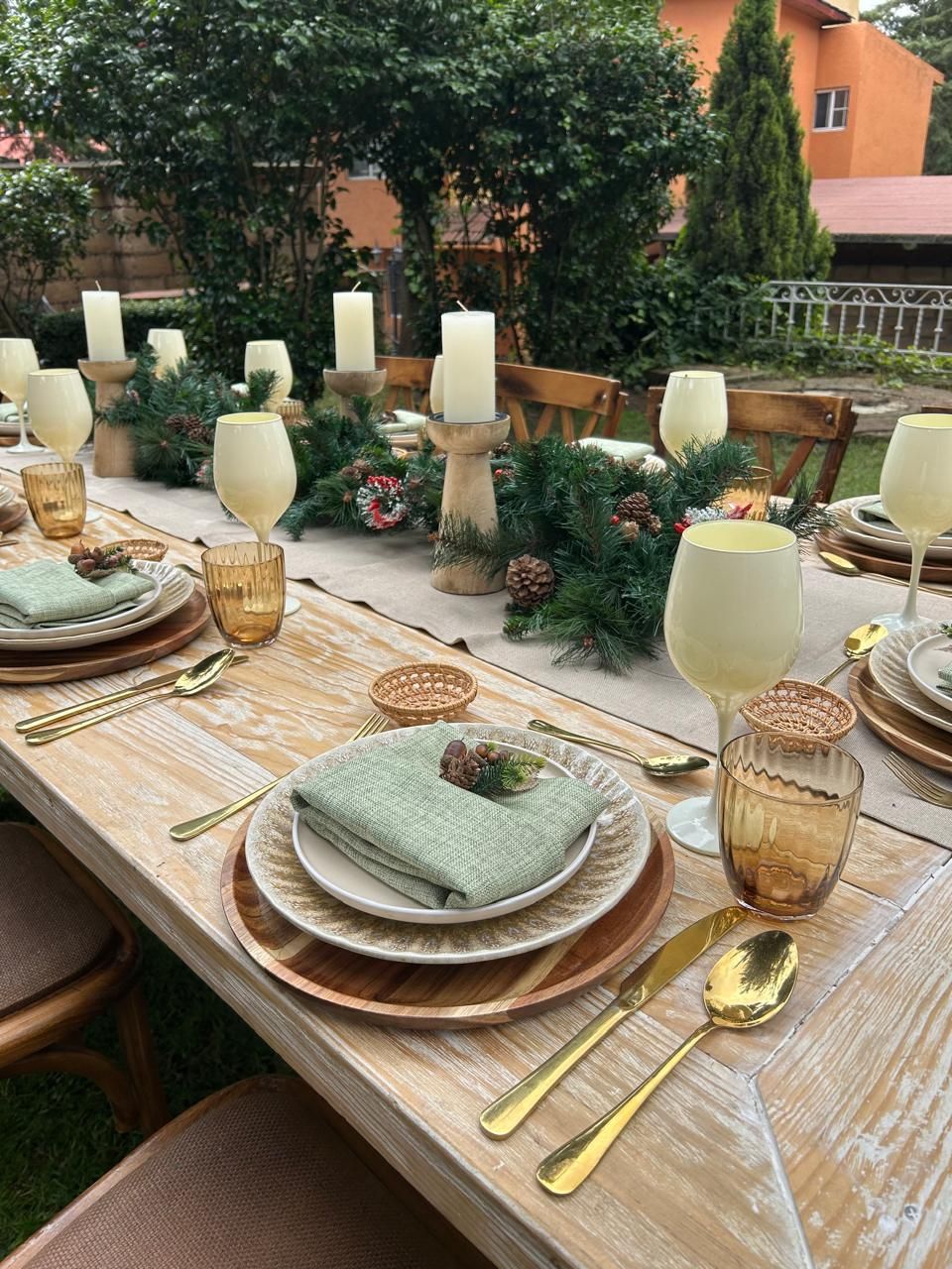 Mesa de comedor al aire libre con cubiertos dorados, velas blancas y servilletas verdes.