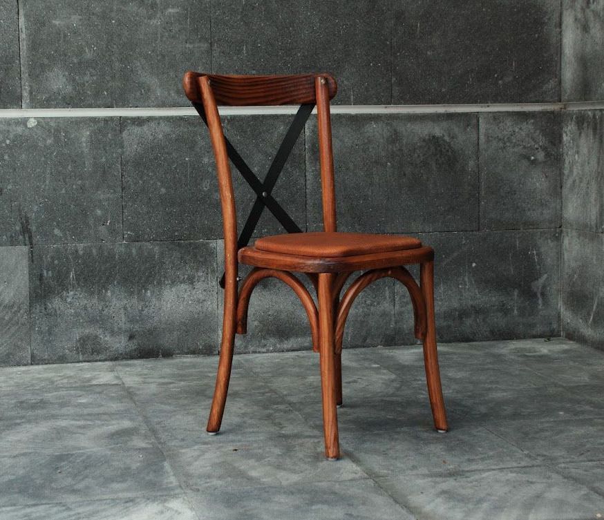 Silla de madera con respaldo en forma de X, asiento y patas de color marrón, colocada contra una pared de piedra gris oscuro.