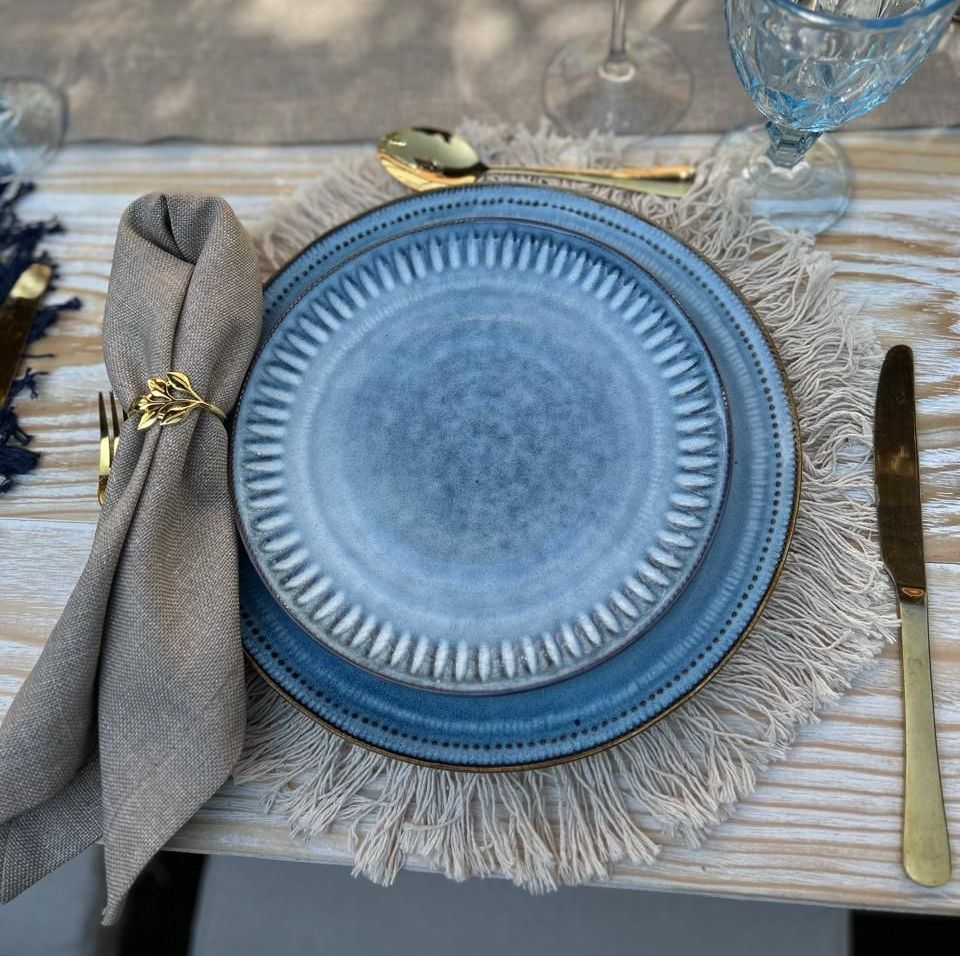 Platos azules sobre una mesa con cubiertos dorados, una servilleta beige y un mantel individual con flecos.