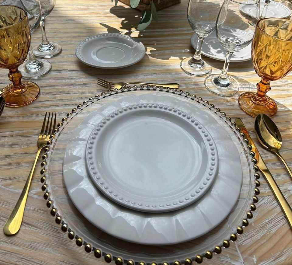 Mesa de comedor formal con cubiertos dorados, vasos color ámbar y platos blancos sobre un plato base con cuentas transparentes.