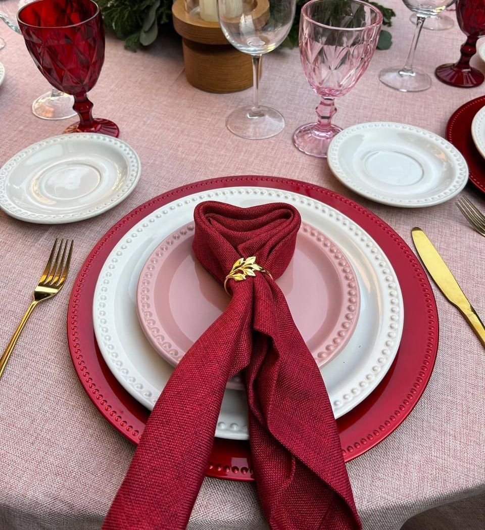 Elegante mesa con platos rojos, blancos y rosas, cubiertos dorados y servilleta en forma de corazón.