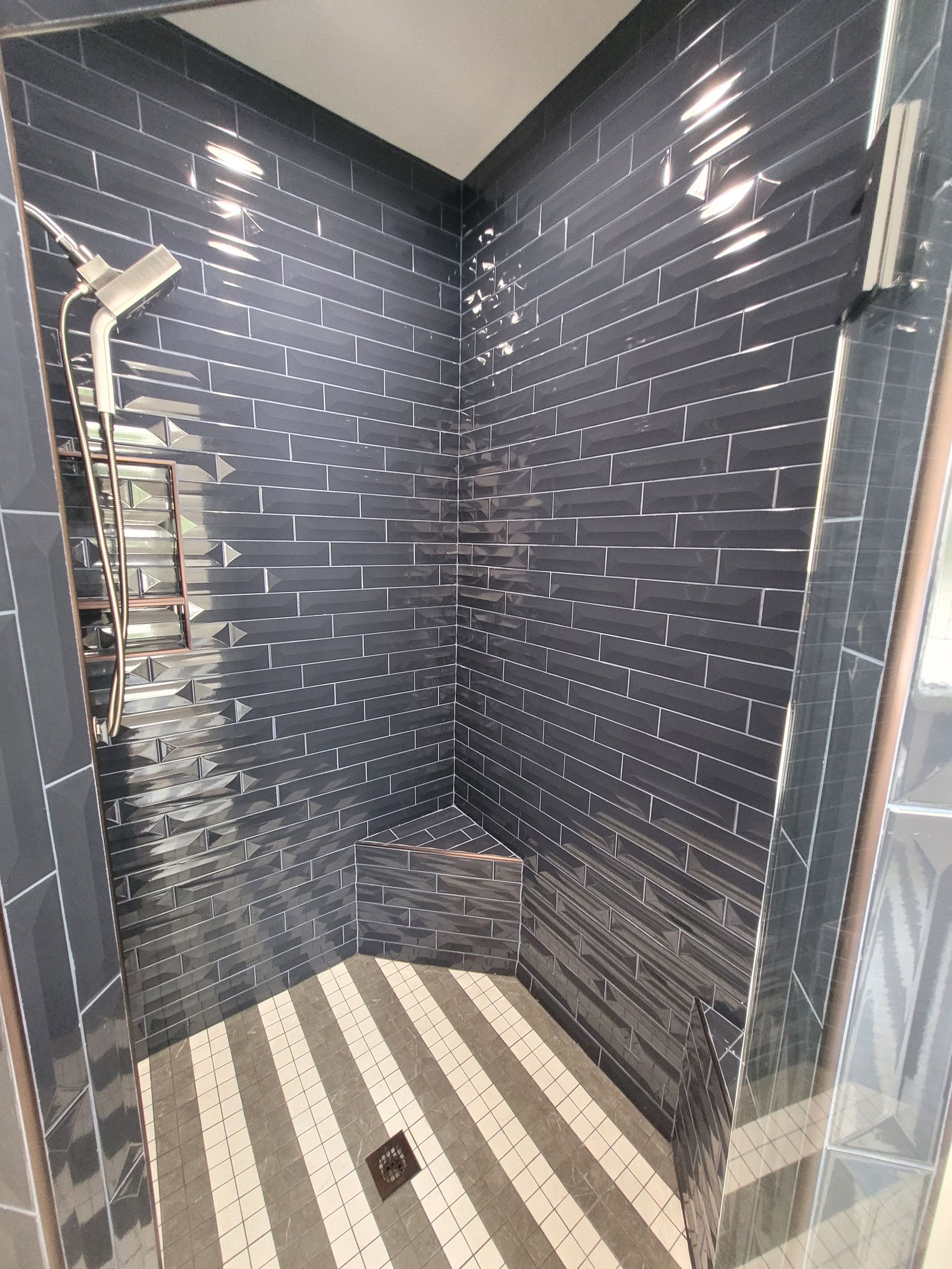 Dark blue tiled shower with gray and white striped floor. Shower head and corner bench are visible.