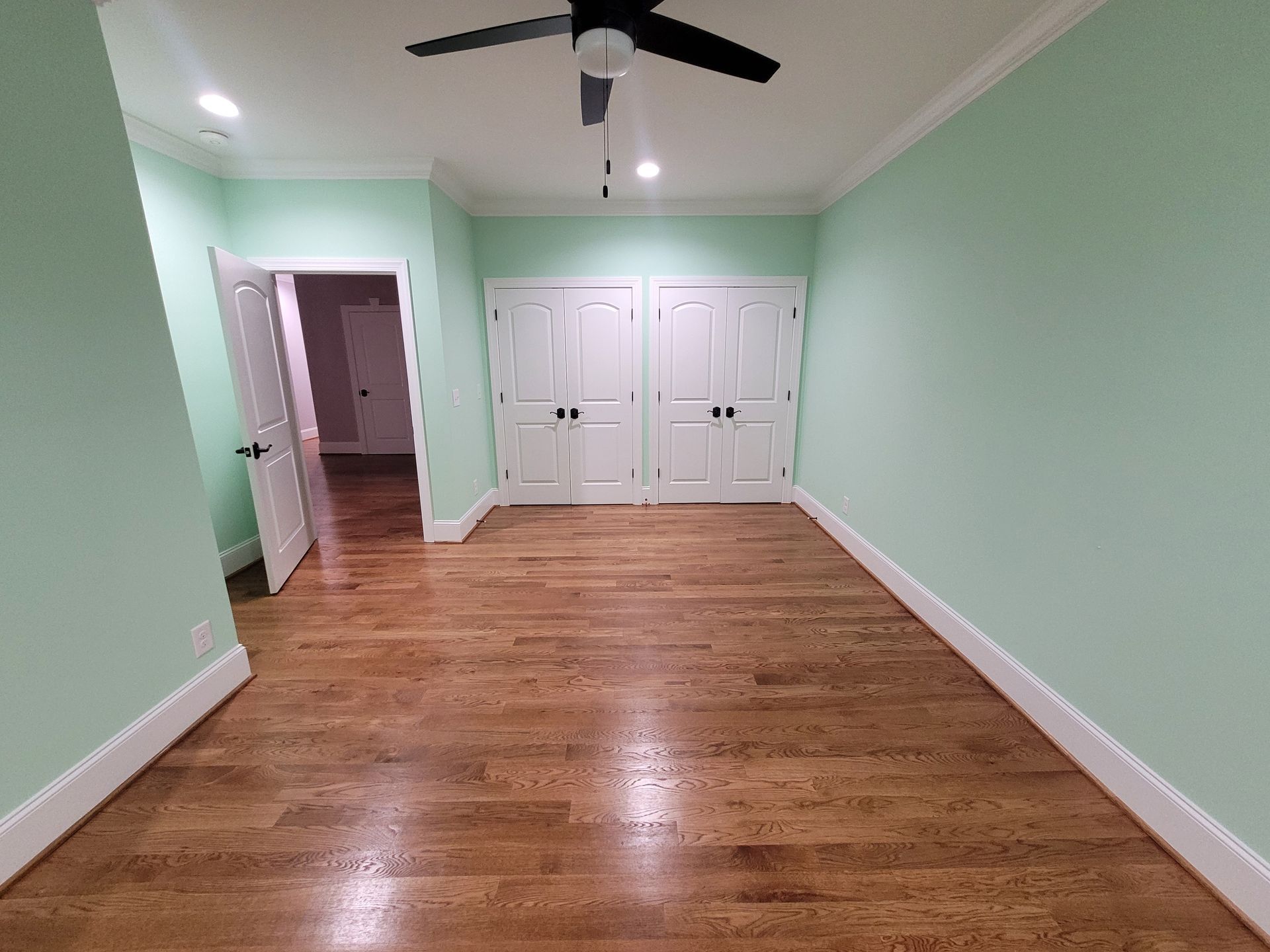 Empty room with mint green walls, hardwood floors, white trim, and closet doors. A doorway leads to another room.