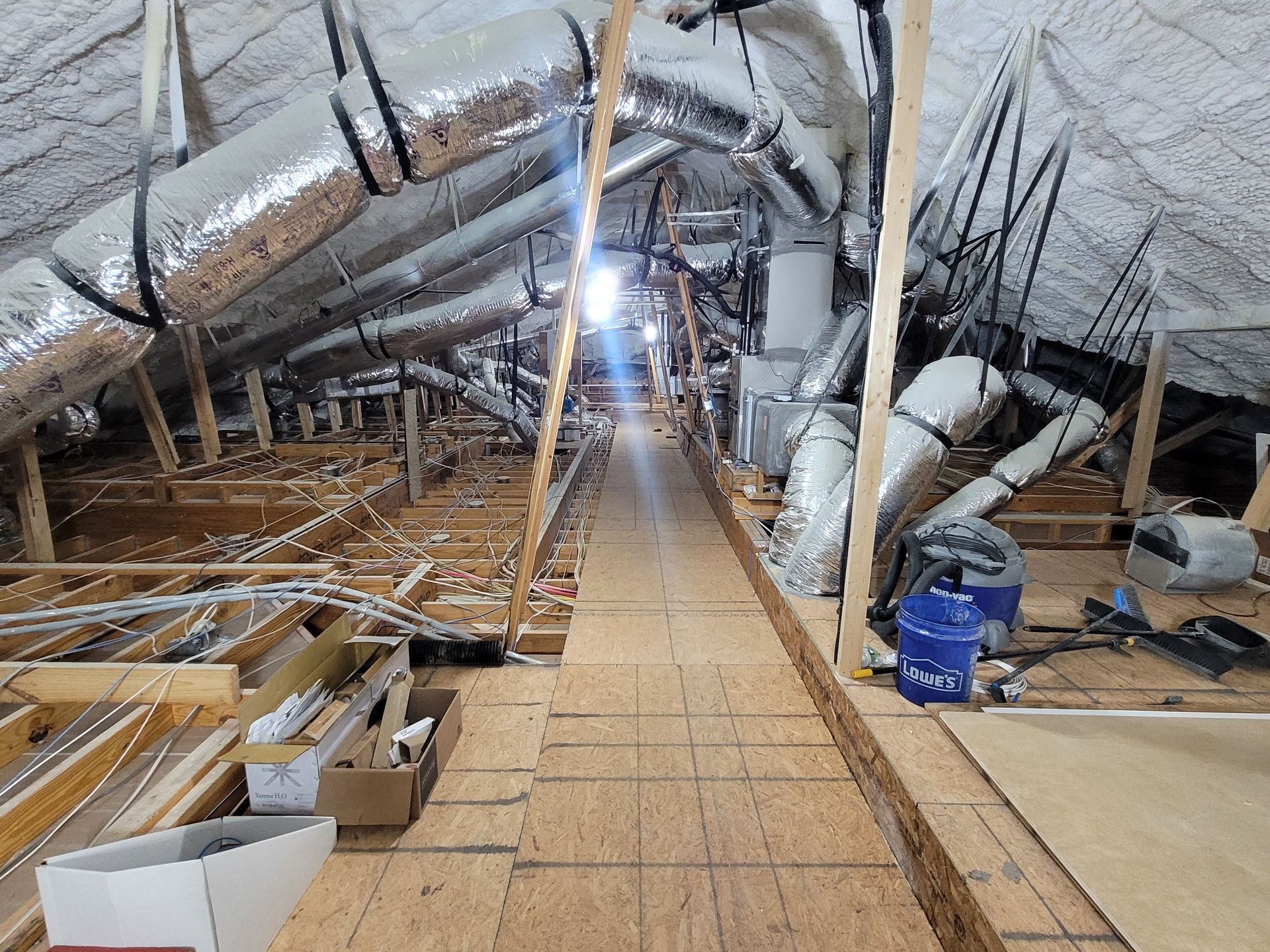 Attic with ductwork and exposed wooden beams; plywood walkway; tools and supplies scattered around.