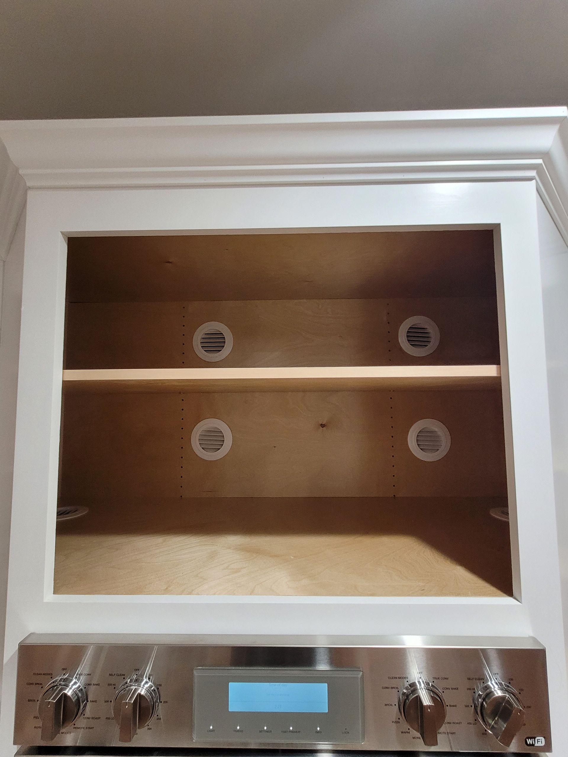 Empty cabinet above a stainless steel range with knob controls; white trim around opening, light wood interior.