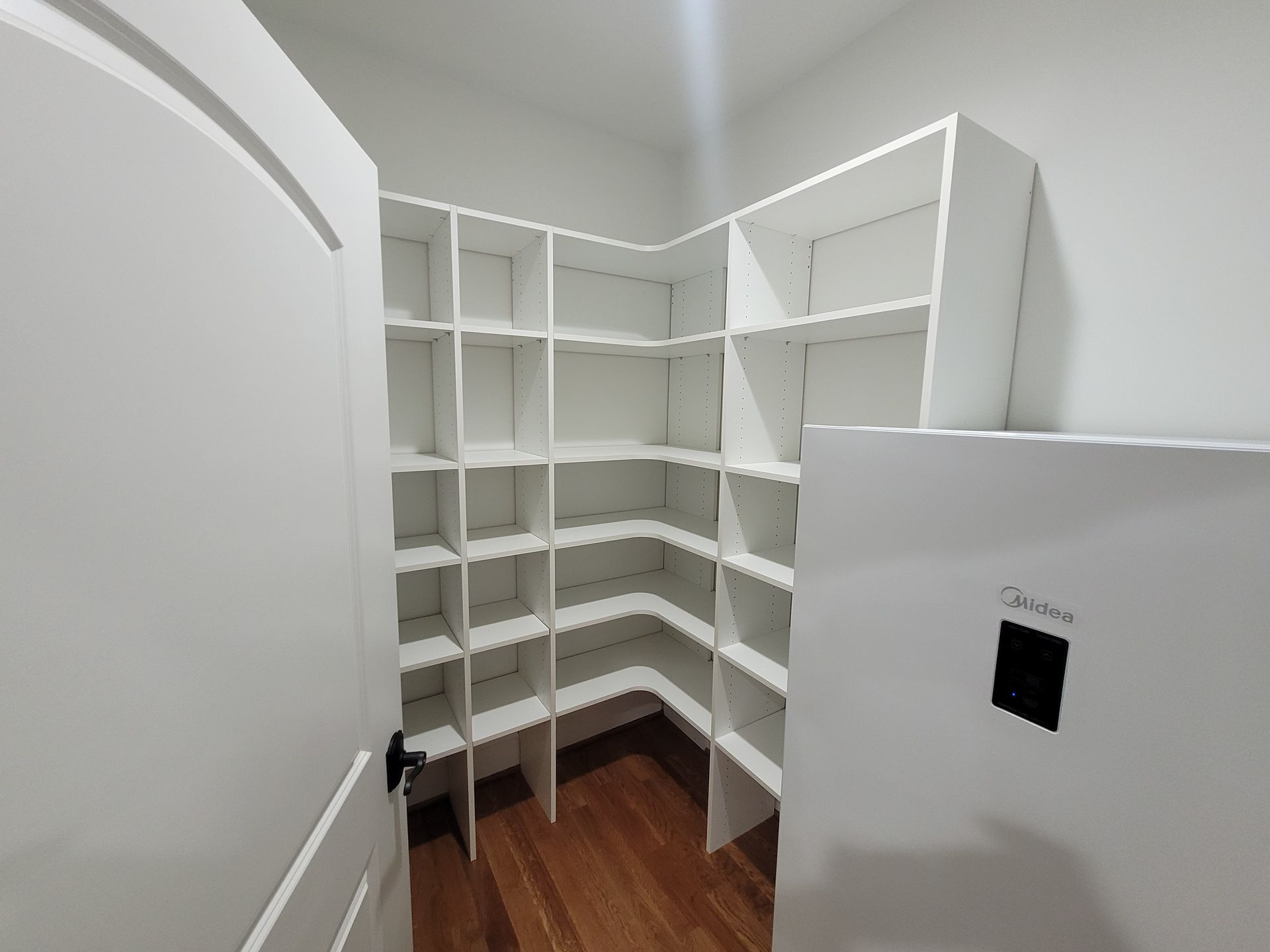 White pantry with built-in shelves in a corner, wooden floor, white door and refrigerator on the right.