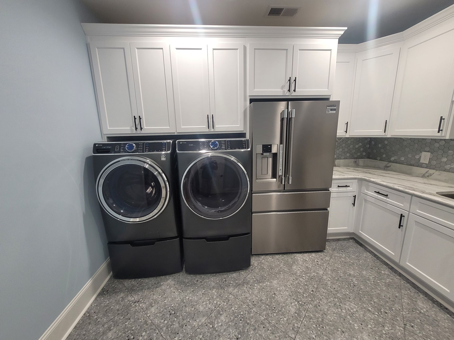 A laundry room with a washer, dryer, refrigerator, white cabinets, and speckled floor.