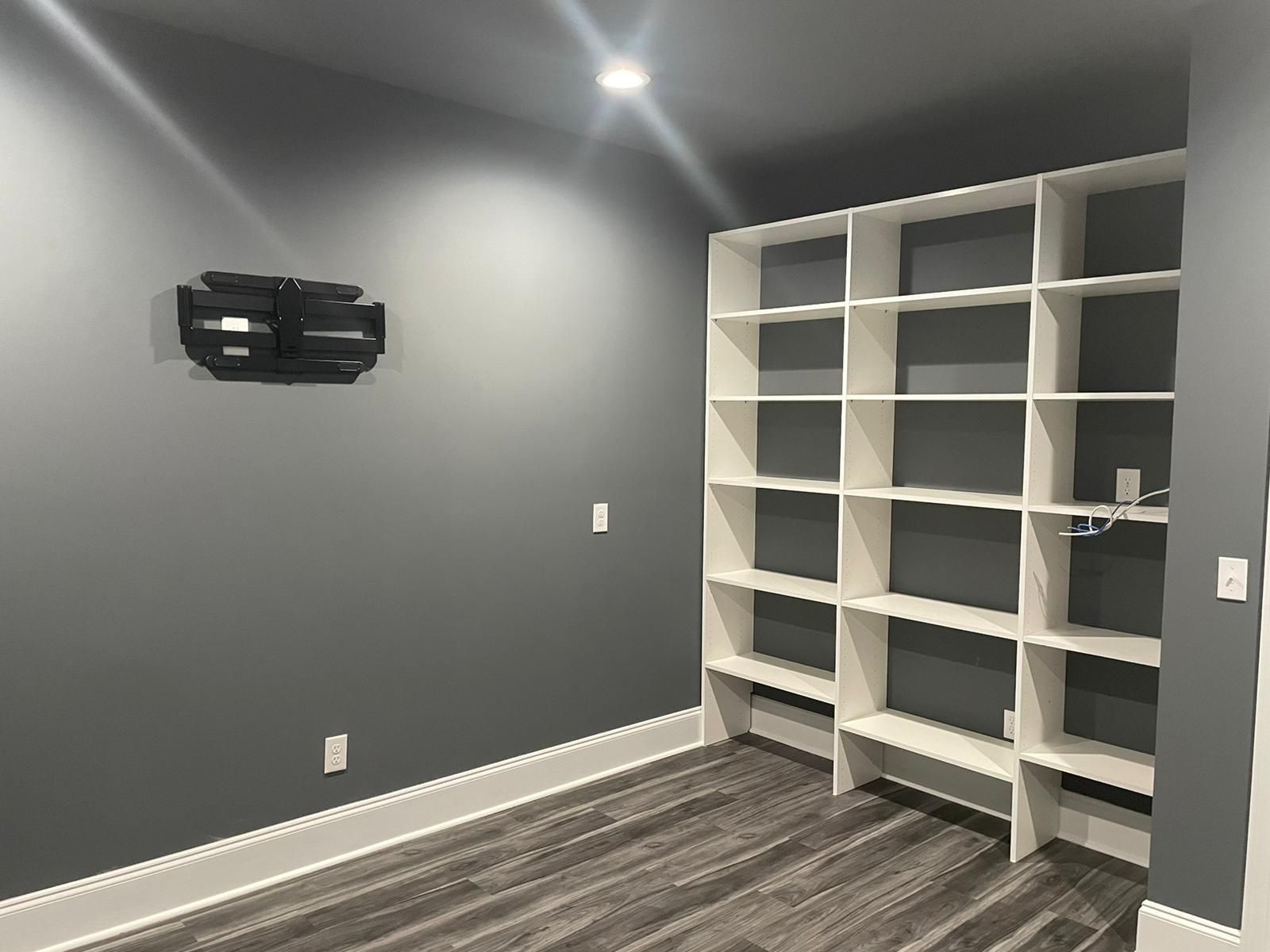 A room with gray walls, white built-in shelves, and wood-look flooring. A projector is mounted on the left.