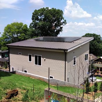 Modern house with angled roof, solar panels, tan siding, surrounded by trees and green grass.