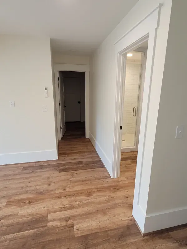 Interior view: hallway with wood floor, white walls, two doorways. One to a bathroom, another to a dark room.