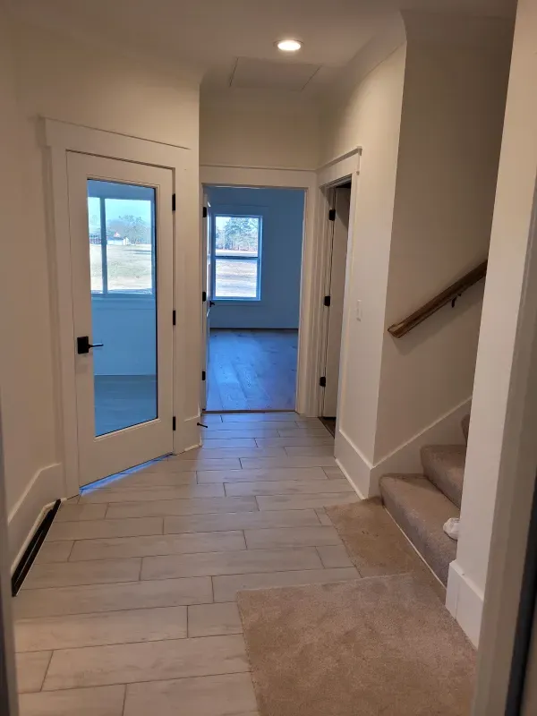 Hallway with tile floor, doors, and stairs. Open door to room with windows.