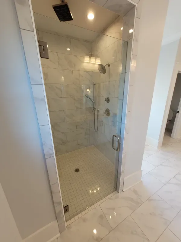 Walk-in shower with glass door, marble walls and floor. Chrome fixtures, white and gray tile, bright interior.