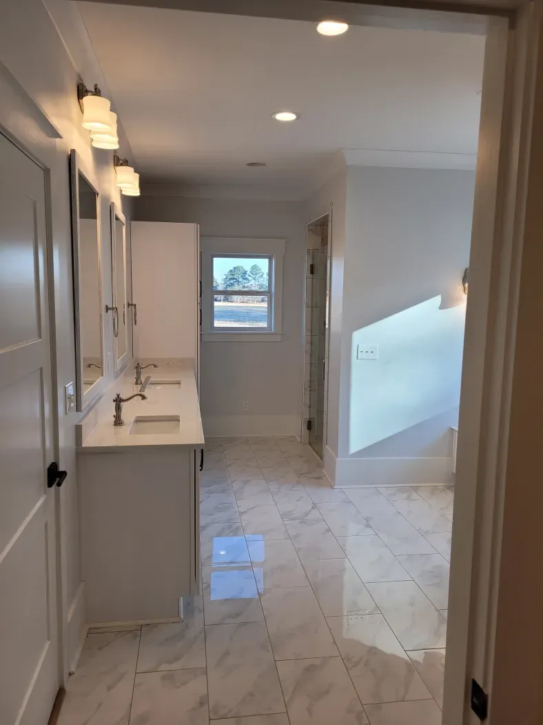 A bright white bathroom with marble floors, vanity, and a window, viewed from the doorway.