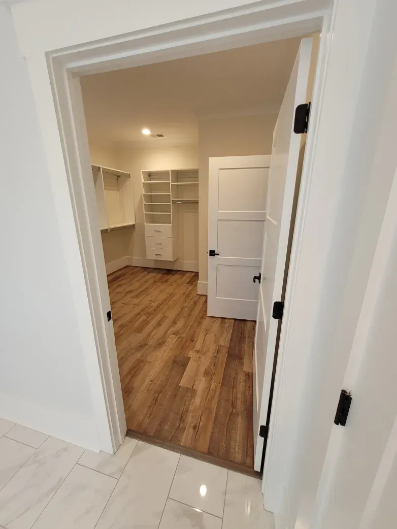 Open doorway to a walk-in closet with built-in shelving and wood flooring; white walls and door, black hardware.