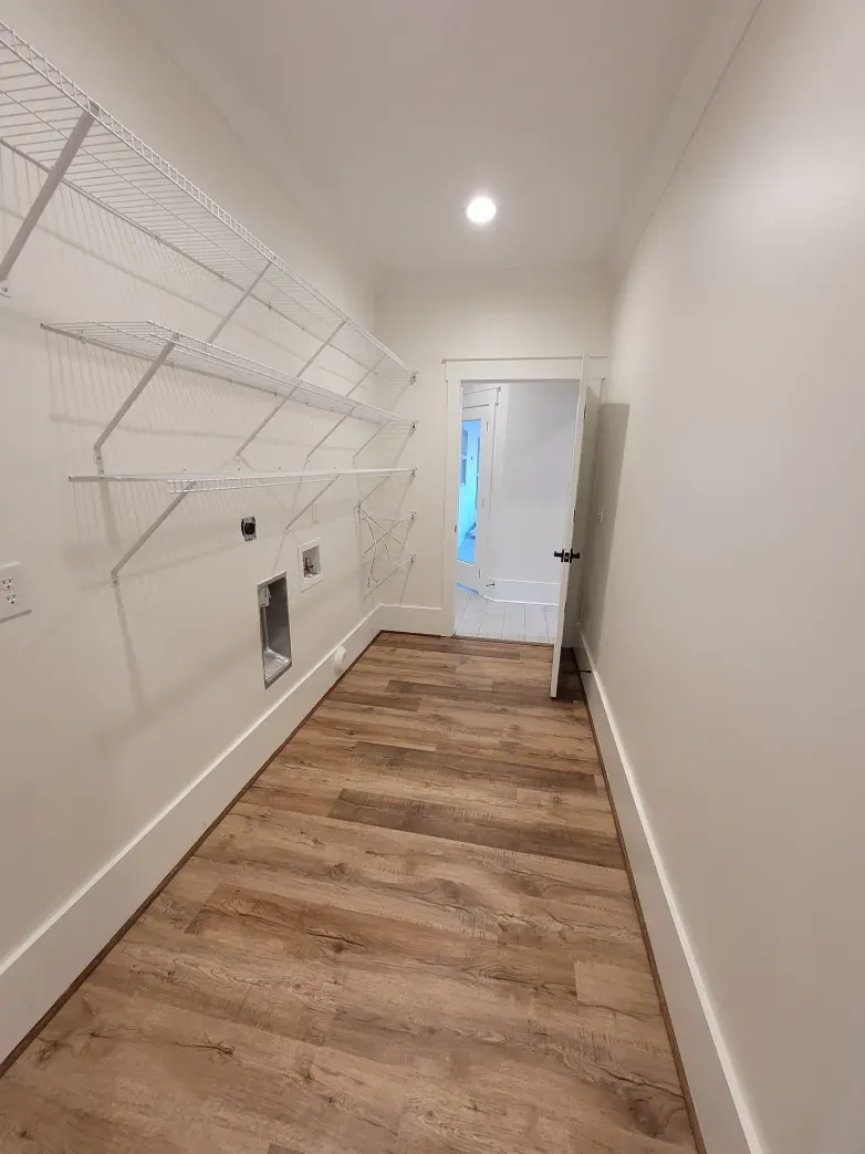 A long, narrow laundry room with wood-look flooring, white walls, and wire shelving. Doorway visible at the end.