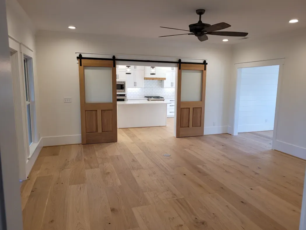 Spacious room with light wood floors, barn doors, and a view of a white kitchen; lit by recessed lights and a ceiling fan.