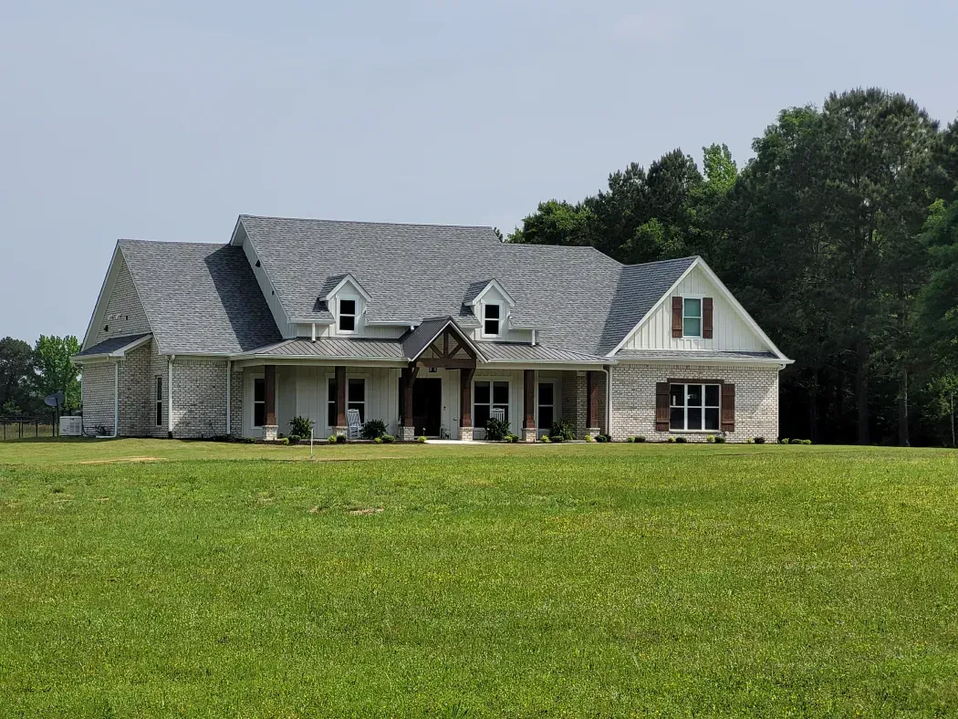 Large, light-colored brick house with gray roof, dormers, and wooden accents on a grassy lawn.
