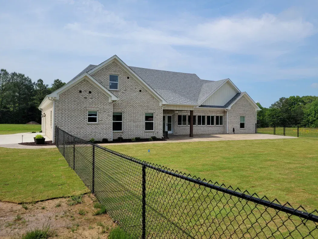 Light brick house with gray roof and black chain-link fence. Green grass and blue sky.