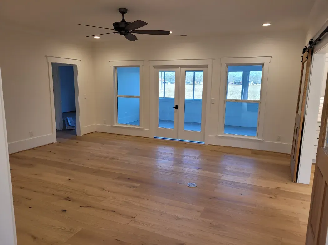 Spacious room with hardwood floors, white walls, and large windows letting in sunlight. A ceiling fan hangs in the center.