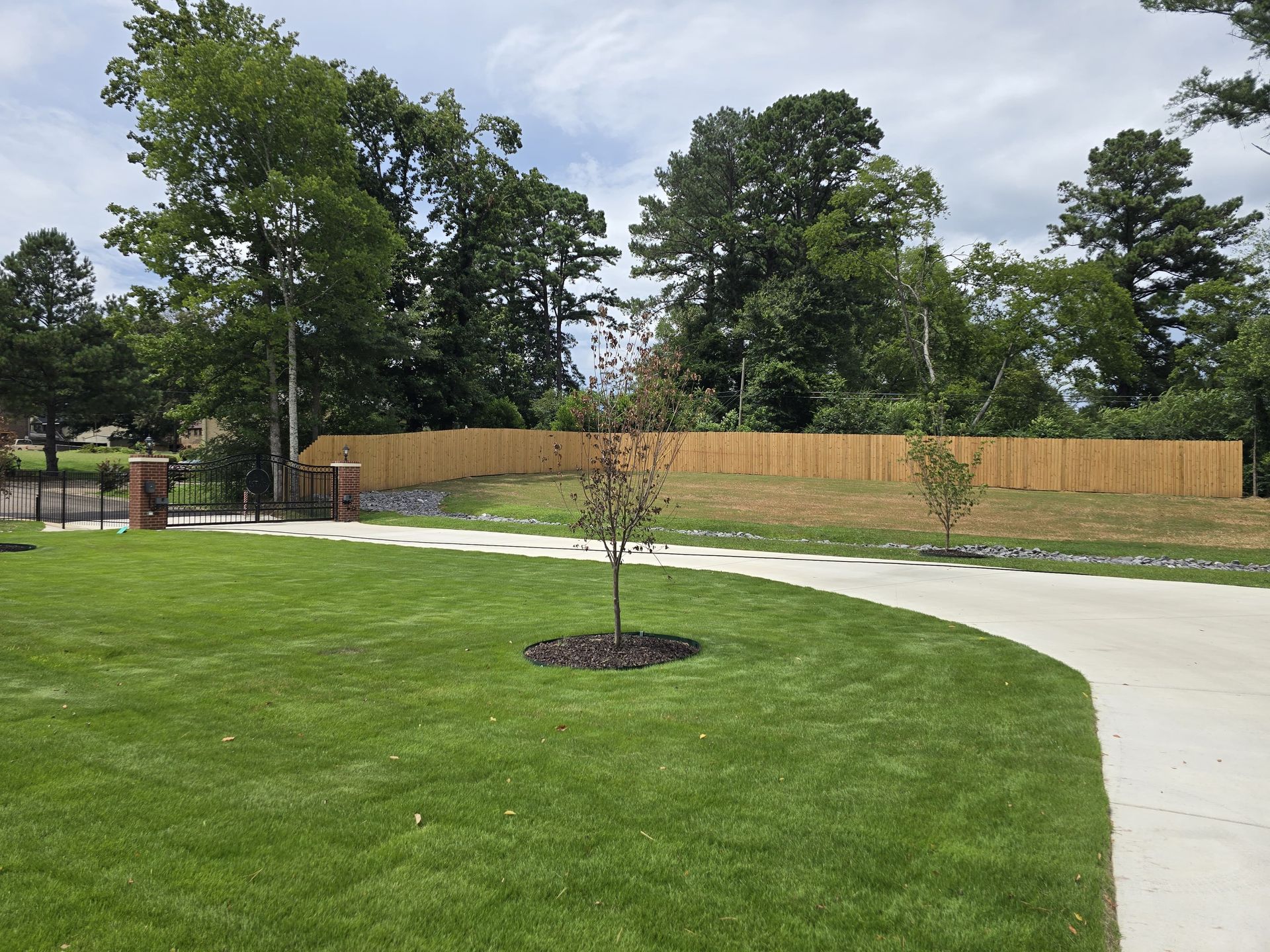 Green lawn with a concrete driveway curving to the right, leading to a wooden fence with trees in the background.