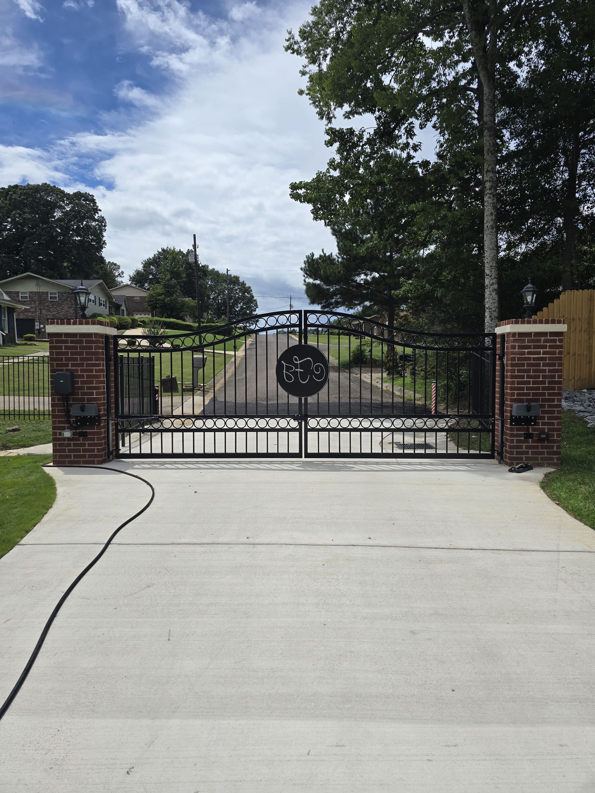 Black wrought-iron driveway gate with a circular emblem between brick pillars on a concrete driveway.