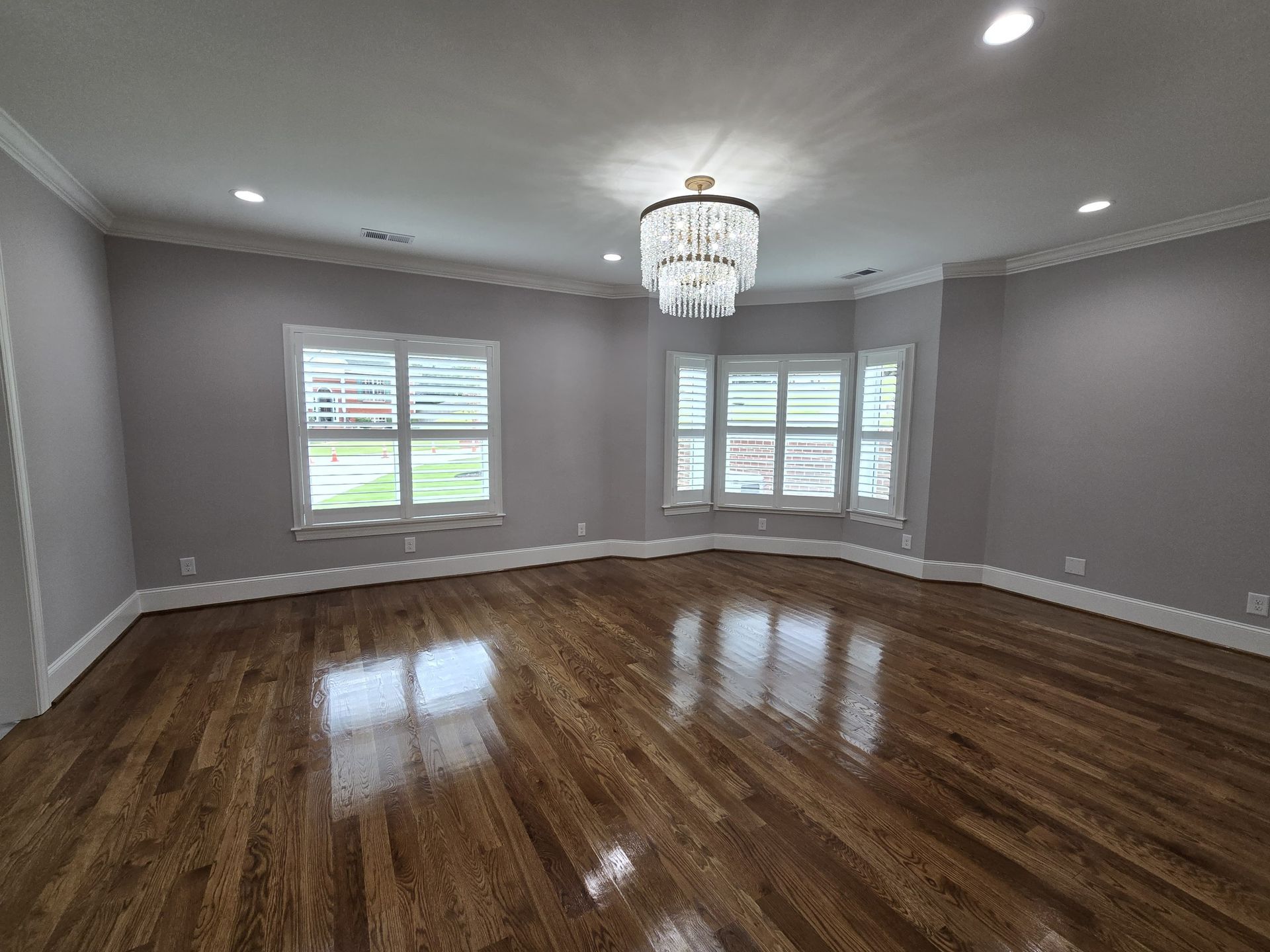 Empty room with hardwood floors, grey walls, and a chandelier. Windows with white shutters.